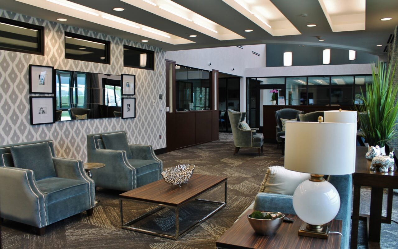 A modern, spacious lounge area with comfortable armchairs, a coffee table, a side table with a lamp, decorative items, and a large plant. The background features a patterned wallpaper, framed pictures, mirrors, and glass panels creating a welcoming atmosphere.