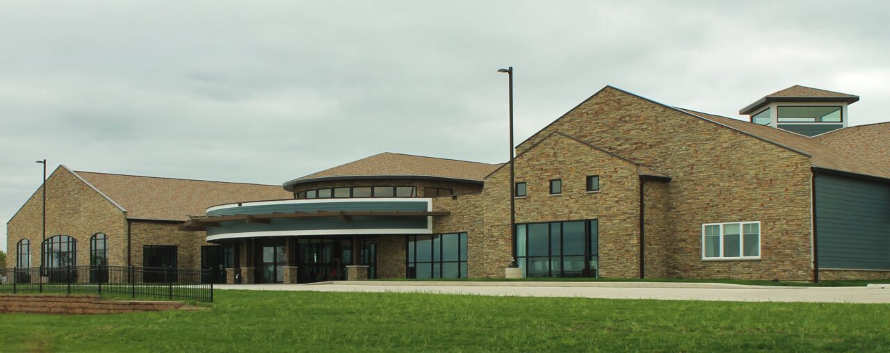 A large, modern rehabilitation center building with a mix of brick and dark gray siding, featuring multiple gabled roofs, large glass windows, and a circular entrance area. The building is surrounded by a grassy lawn and a paved walkway, with a cloudy sky overhead.