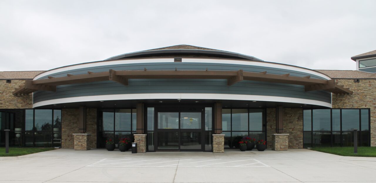 Front view of a modern rehabilitation center building with large glass doors, stone columns, and a curved roof. There are potted plants with red flowers near the entrance.