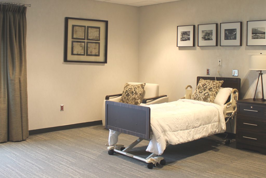 A hospital room with a bed on wheels, white bedding, and two patterned pillows. There is a dark wooden nightstand with a lamp and a small decorative object. A beige armchair with a patterned pillow is next to the bed. Framed black-and-white photographs hang on the wall, and a window with closed curtains is on the left.