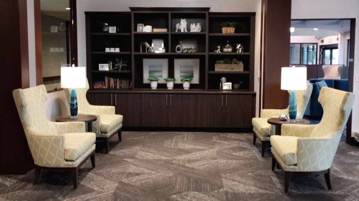 A cozy seating area with four yellow armchairs, each paired with a small wooden side table and a white table lamp. In the background, there is a dark wooden shelving unit with decorative items, books, and potted plants. The area has a modern, welcoming atmosphere.