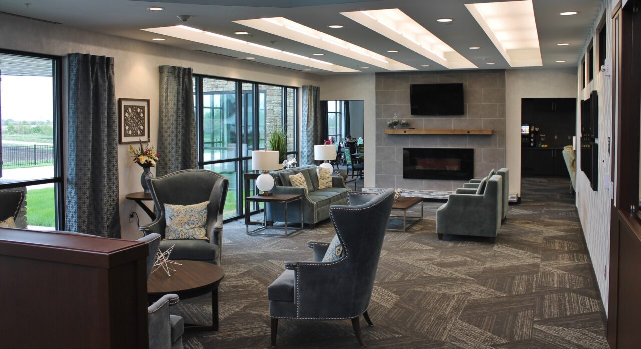A modern, well-lit lounge area with large windows, gray armchairs, a sofa, and a fireplace with a mounted TV above it. There are side tables with lamps and decorative pillows on the furniture. The space features a mix of natural and artificial lighting, with curtains and a view of a green outdoor area.