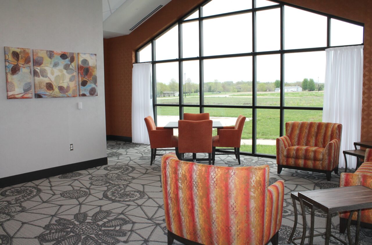 A spacious room with large, floor-to-ceiling windows showing a green outdoor landscape. The room has orange upholstered chairs around a table, a patterned carpet, and a white wall with colorful abstract artwork. White curtains are partially drawn to the sides of the windows.