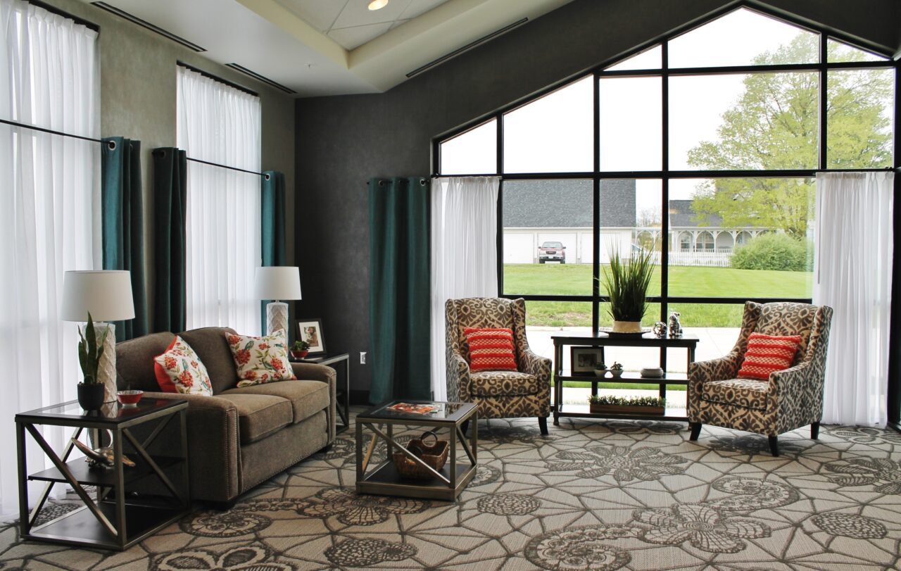 A bright, modern living room with large windows and white curtains, featuring a beige sofa with floral pillows, two patterned armchairs with red pillows, a glass coffee table, and side tables with lamps, overlooking a green lawn and trees outside.