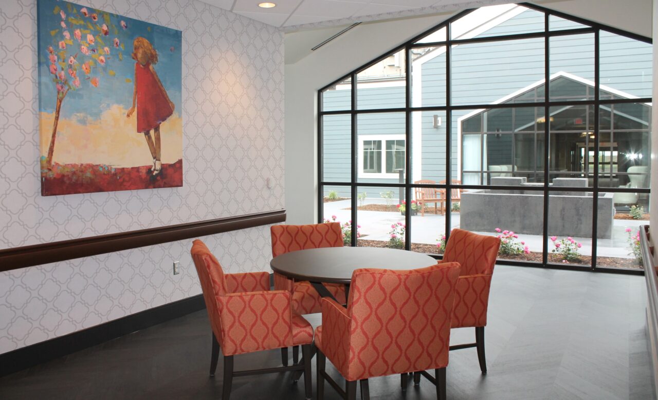 A cozy seating area with four orange patterned chairs arranged around a round table, set against a wall with a colorful painting of a girl in a red dress walking near a tree with pink flowers. Large glass windows reveal an outdoor patio with flower beds and outdoor seating.