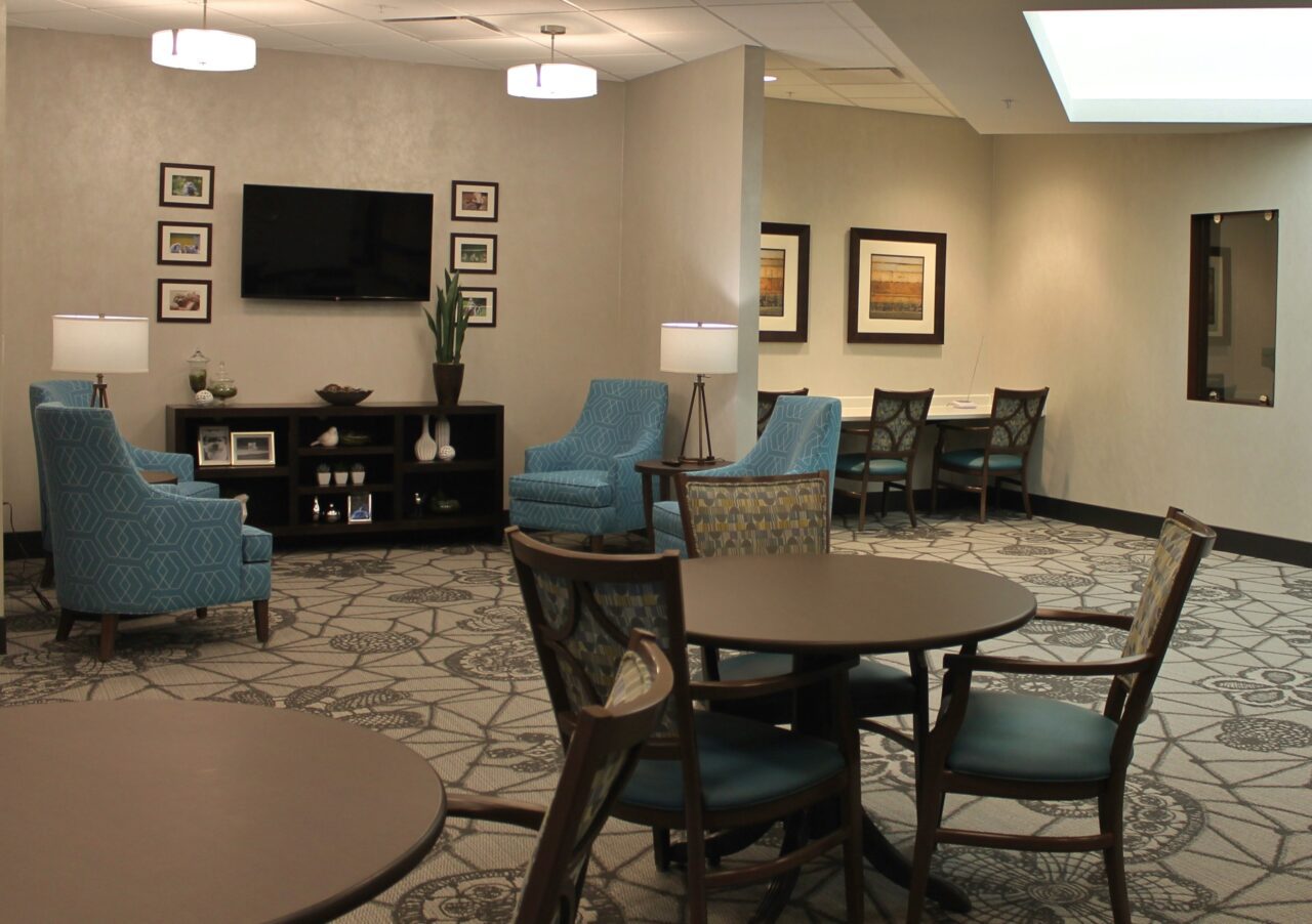 A cozy lounge area with round wooden tables and chairs, two blue patterned armchairs with small lamps, a black shelf with decorative items and a tall potted plant, a wall-mounted TV surrounded by framed photos, and a small dining table with four chairs against the wall. The room has patterned carpet flooring and soft lighting.