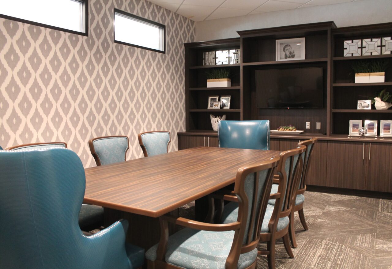 A conference room with a wooden table surrounded by six chairs, some with blue upholstery and others with patterned fabric. The room has a patterned wallpapered wall, three narrow windows near the ceiling, and a large dark wooden cabinet with shelves, decorative items, framed photos, and a flat-screen TV.