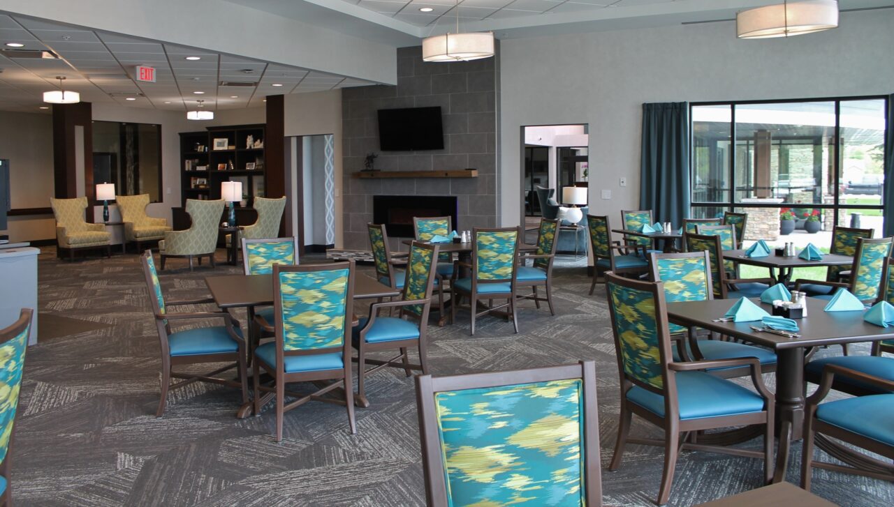 A spacious dining area with several round and rectangular tables set with turquoise napkins and silverware. The chairs have blue cushions and patterned fabric backs. Large windows with blue curtains allow natural light, and there is a fireplace with a TV mounted above on a gray wall. In the background, there are armchairs and bookshelves.