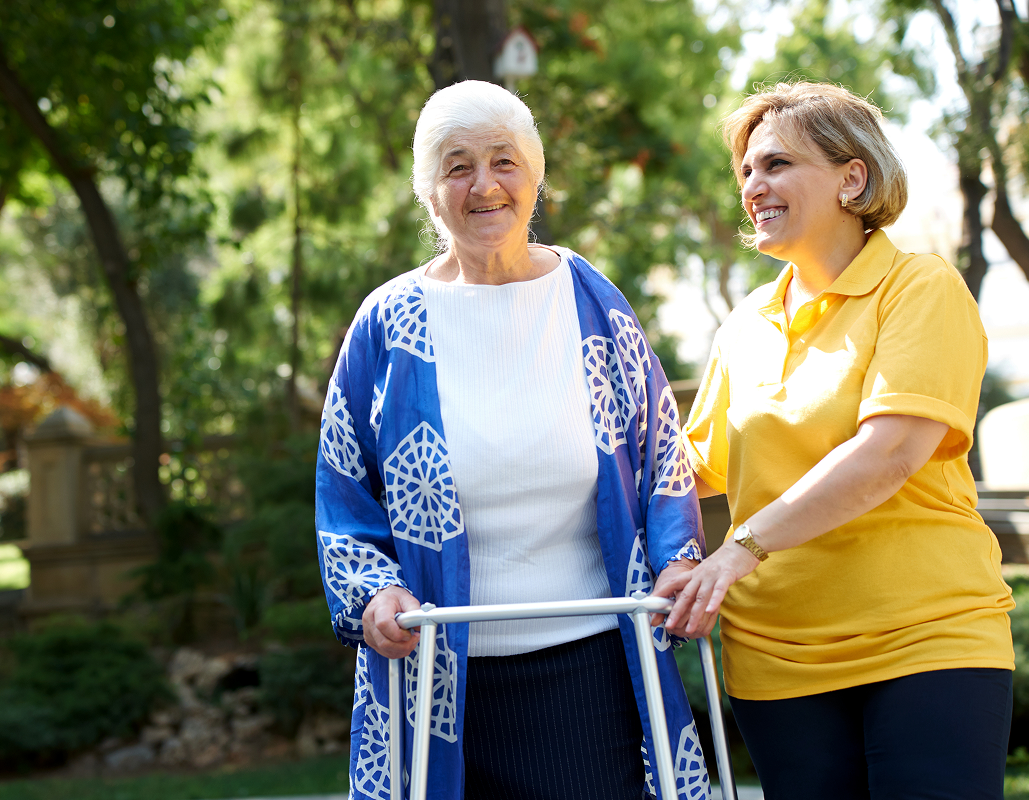 An elderly woman with gray hair using a walker, smiling, standing outdoors in a park-like setting, assisted by a smiling woman in a yellow shirt holding her arm.